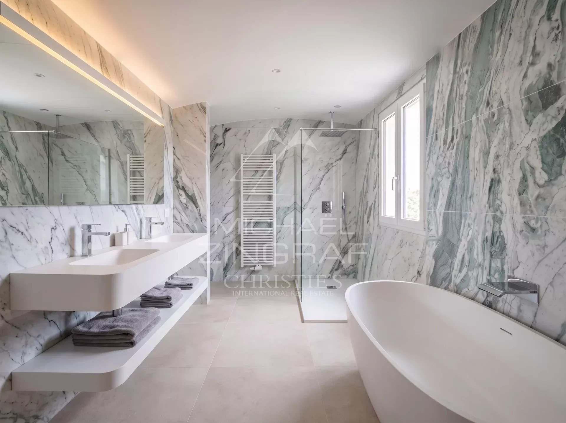 Salle de bain moderne avec baignoire et douche, murs en marbre, fenêtres laissant entrer la lumière.