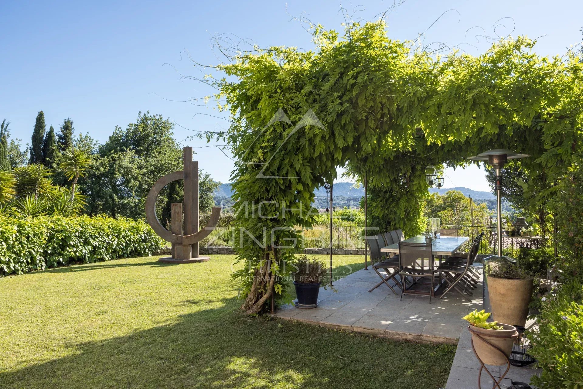 Terrasse d'un mas provençal avec vue sur les collines, entourée de verdure et d'une sculpture moderne.