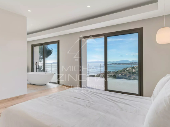 Bright, minimalist bedroom with white bedding and a freestanding bathtub beside large glass doors opening to a balcony with an ocean view.