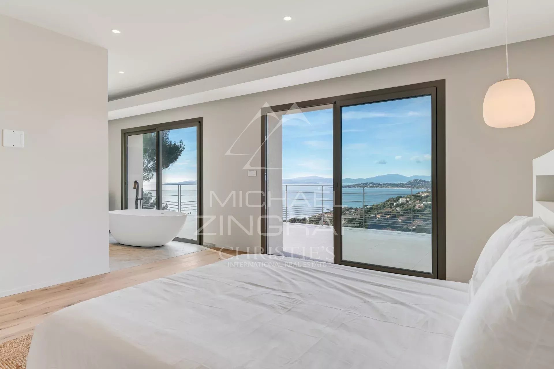 Bright, minimalist bedroom with white bedding and a freestanding bathtub beside large glass doors opening to a balcony with an ocean view.