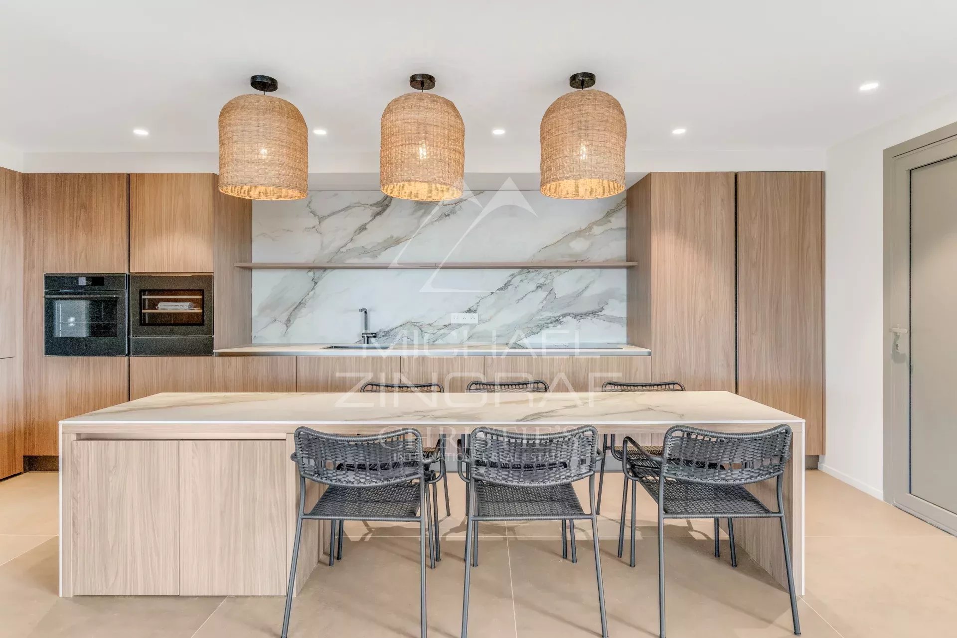 Modern kitchen with a marble backsplash, wooden cabinets, and a light-colored island; three woven pendant lights above and black wire chairs along the island.