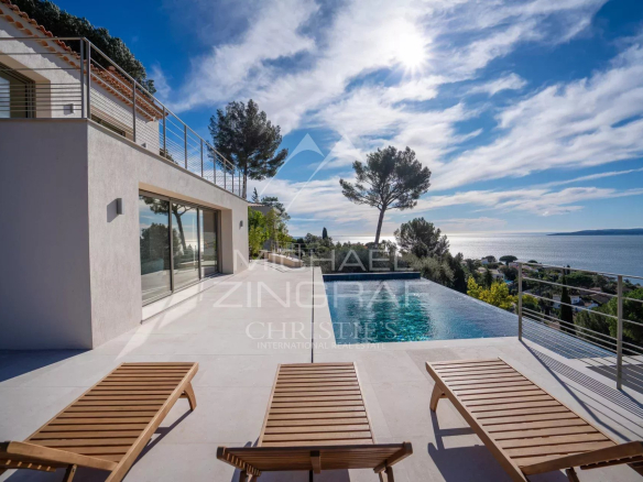 Modern villa terrace with an infinity pool looking toward the sea, wooden sun loungers edged by a glass railing under a sunny sky.