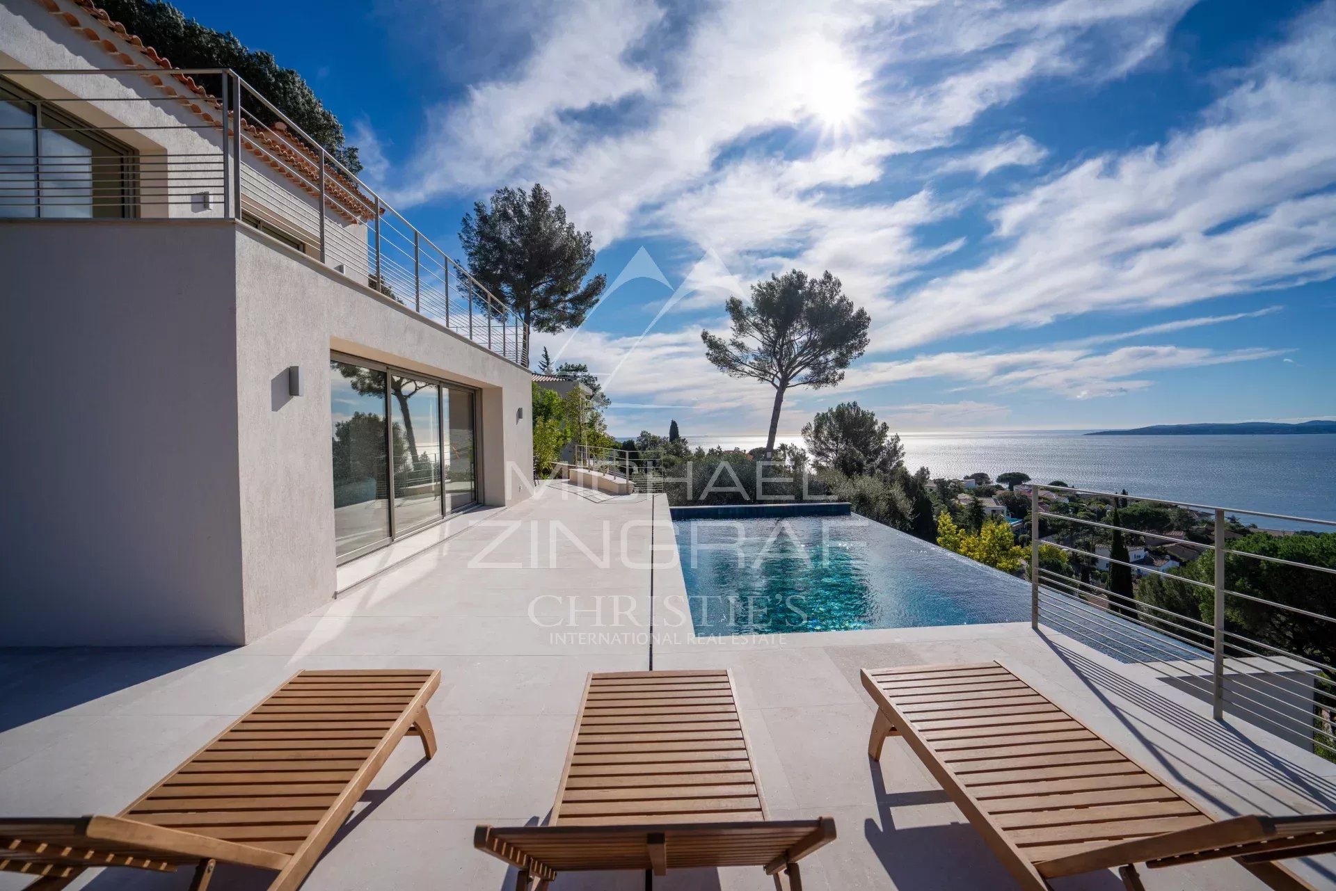 Modern villa terrace with an infinity pool looking toward the sea, wooden sun loungers edged by a glass railing under a sunny sky.