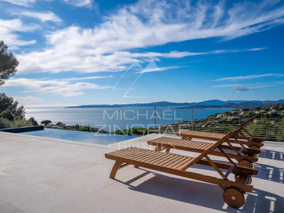 Terrace with an infinity pool and three wooden sun loungers facing a blue sea and distant hills.