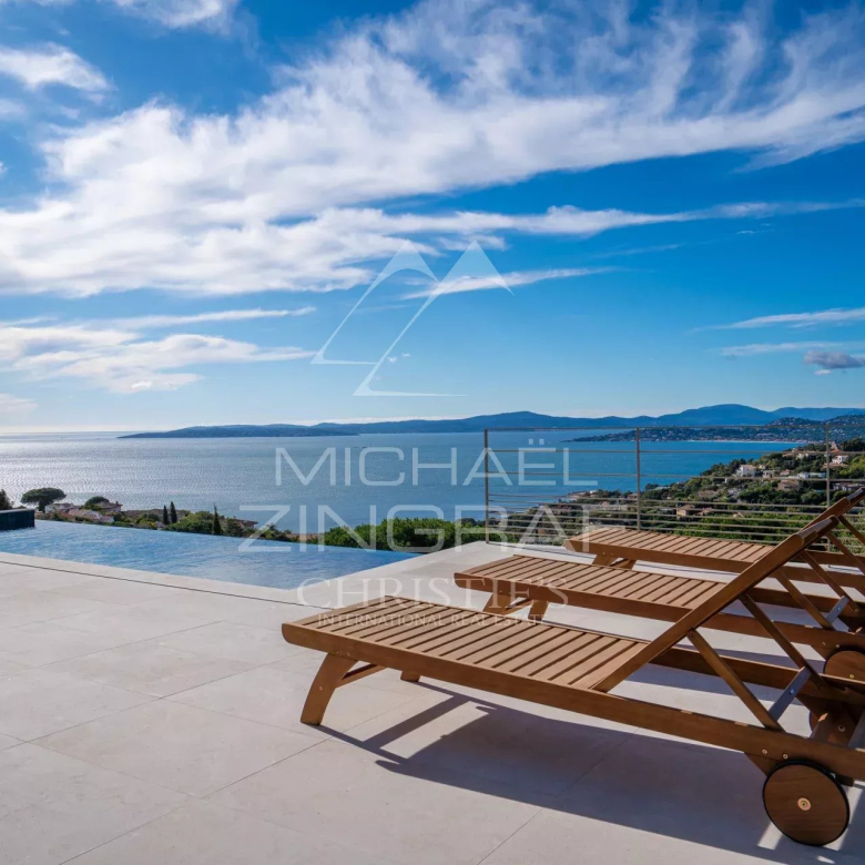 Terrace with an infinity pool and three wooden sun loungers facing a blue sea and distant hills.
