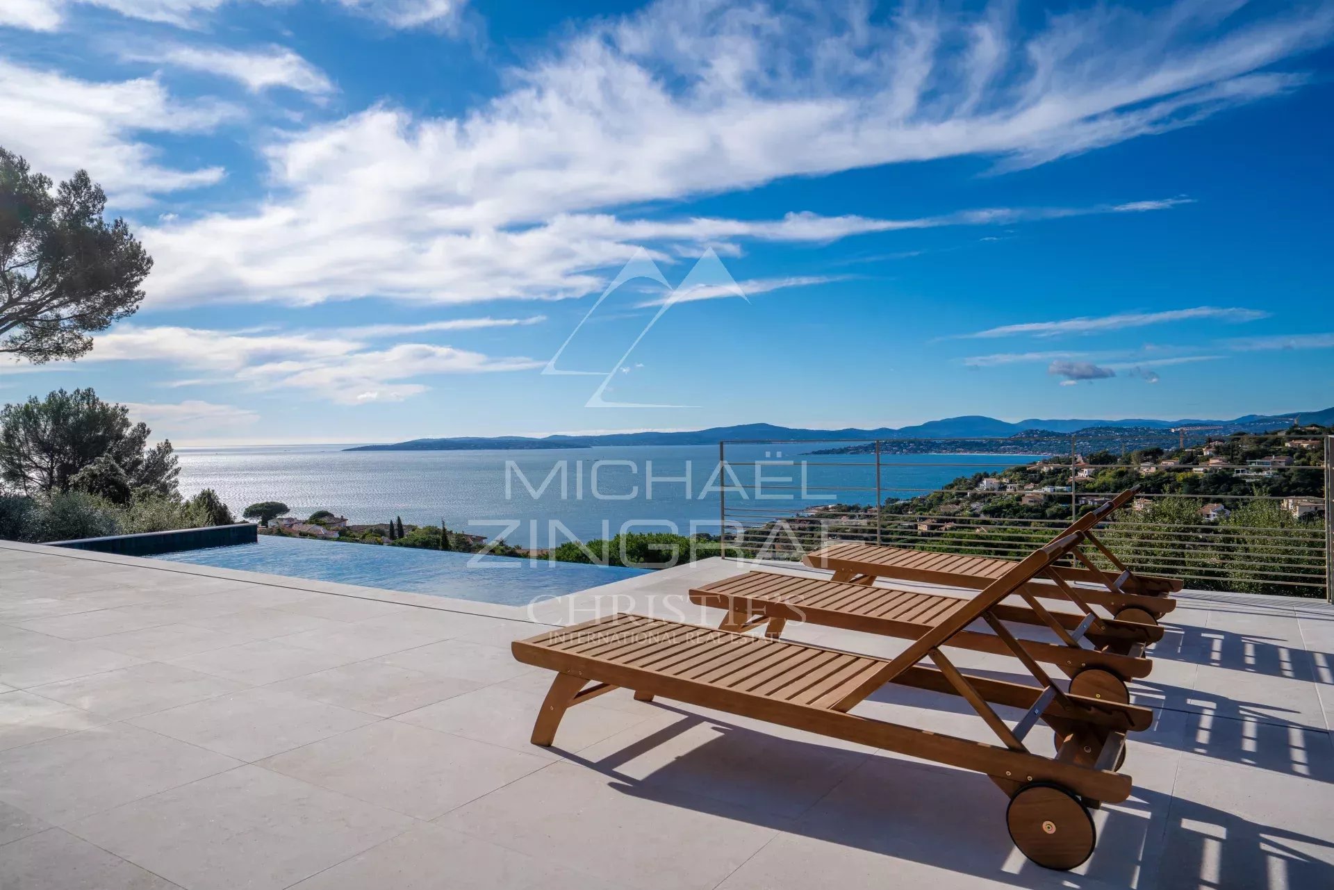 Terrace with an infinity pool and three wooden sun loungers facing a blue sea and distant hills.