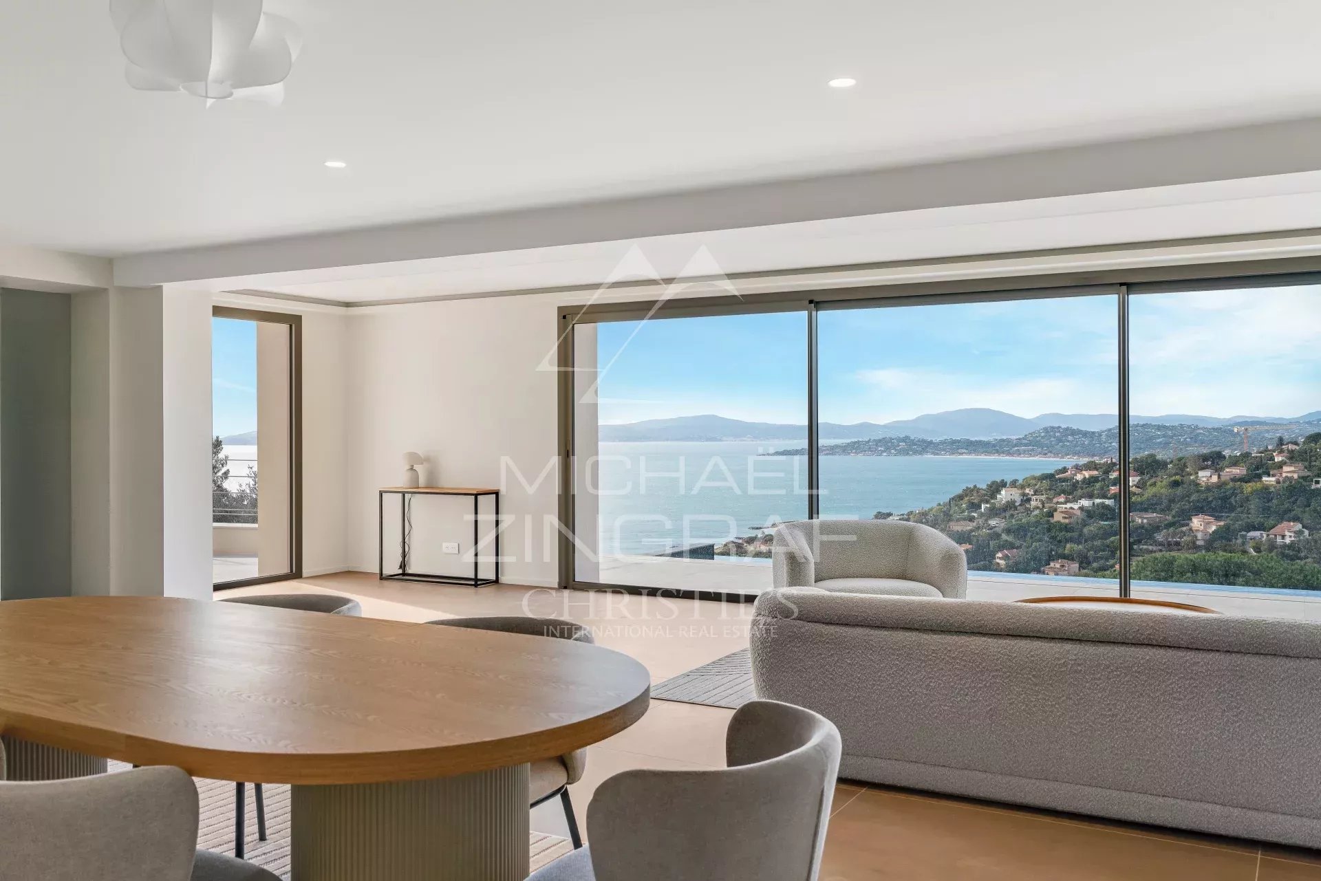 Spacious living area with a round wooden dining table and gray chairs, and large windows offering a coastal panorama.