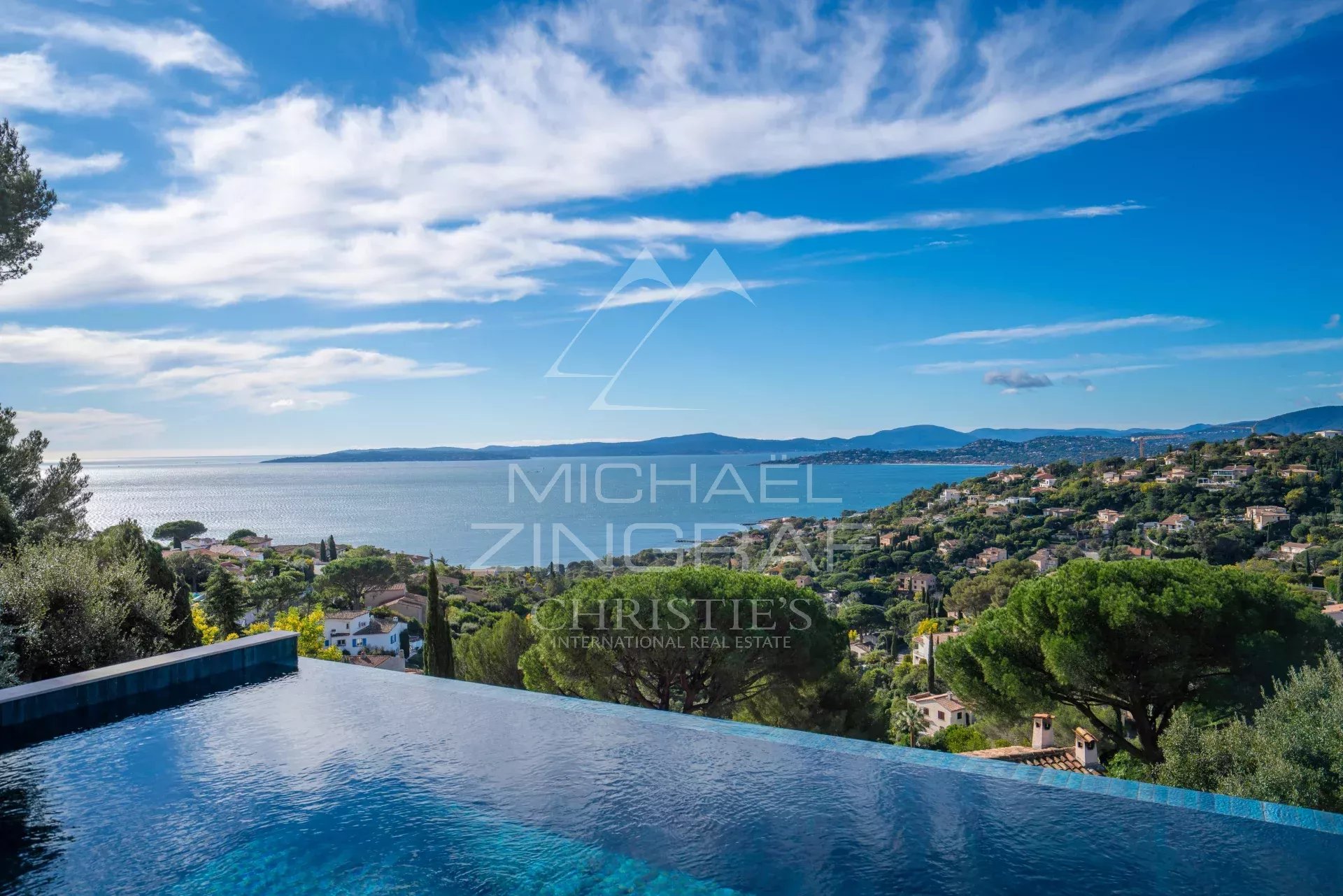 Infinity pool in the foreground with a coastal town, sea, and blue sky beyond.