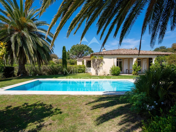 A single-story Mediterranean house with a tiled roof and covered veranda, set behind a rectangular swimming pool amid lush green lawn and palm trees under a bright blue sky.