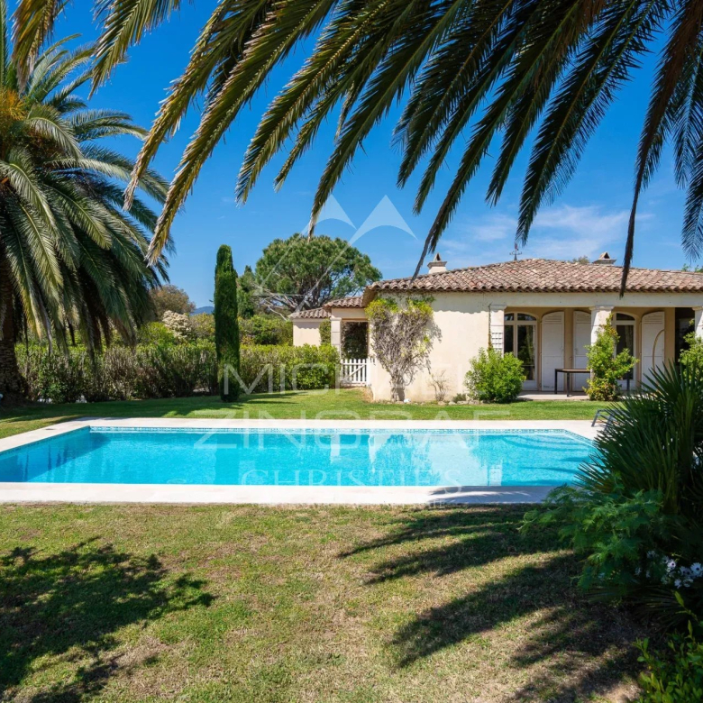 A single-story Mediterranean house with a tiled roof and covered veranda, set behind a rectangular swimming pool amid lush green lawn and palm trees under a bright blue sky.