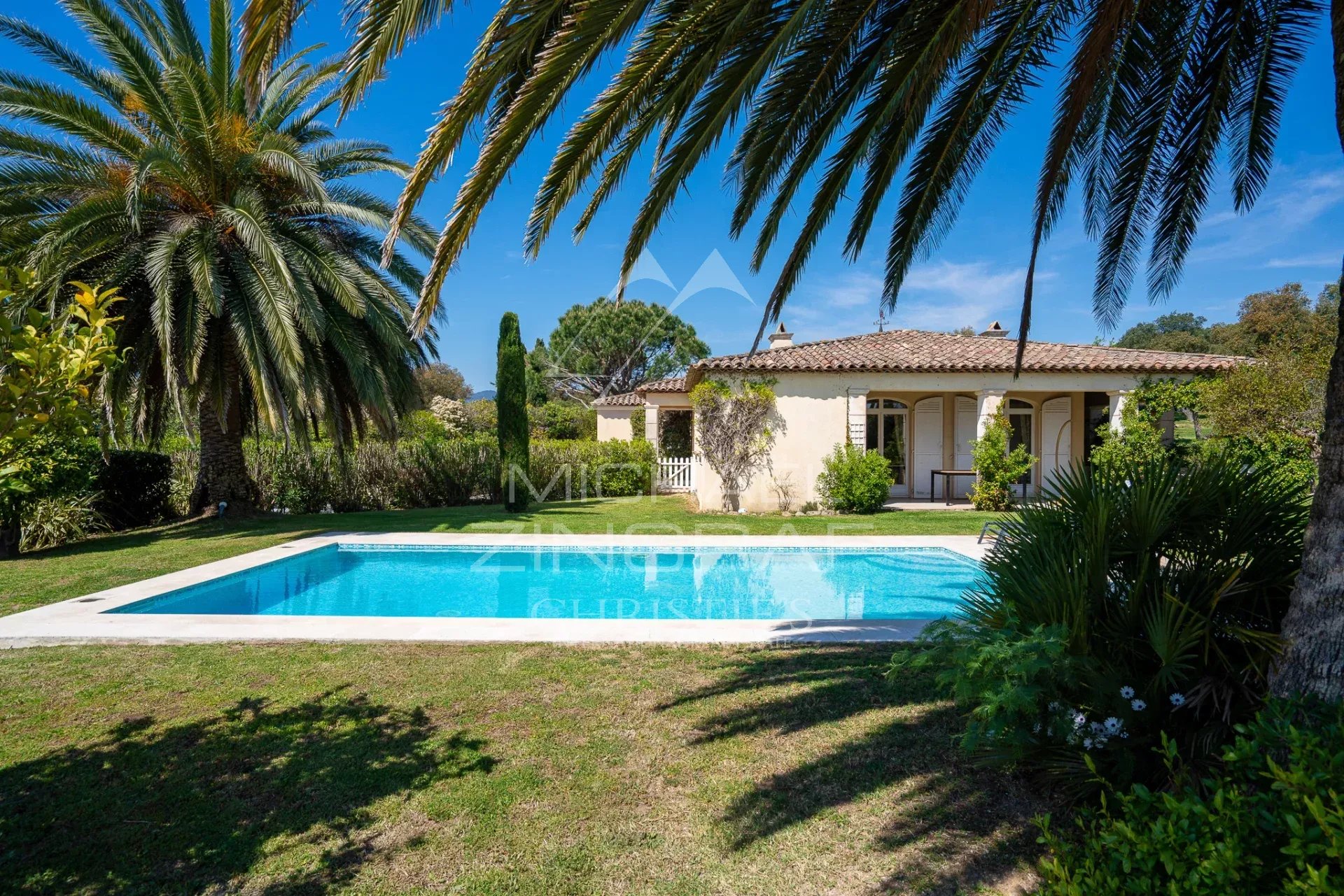 A single-story Mediterranean house with a tiled roof and covered veranda, set behind a rectangular swimming pool amid lush green lawn and palm trees under a bright blue sky.