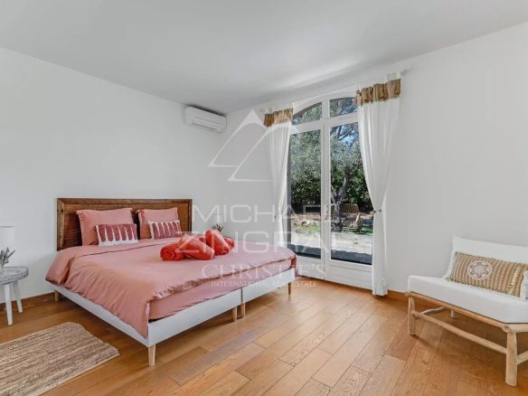 Bright bedroom with a pink-beddinged bed, wooden headboard, white walls, and glass doors opening to a garden outside.