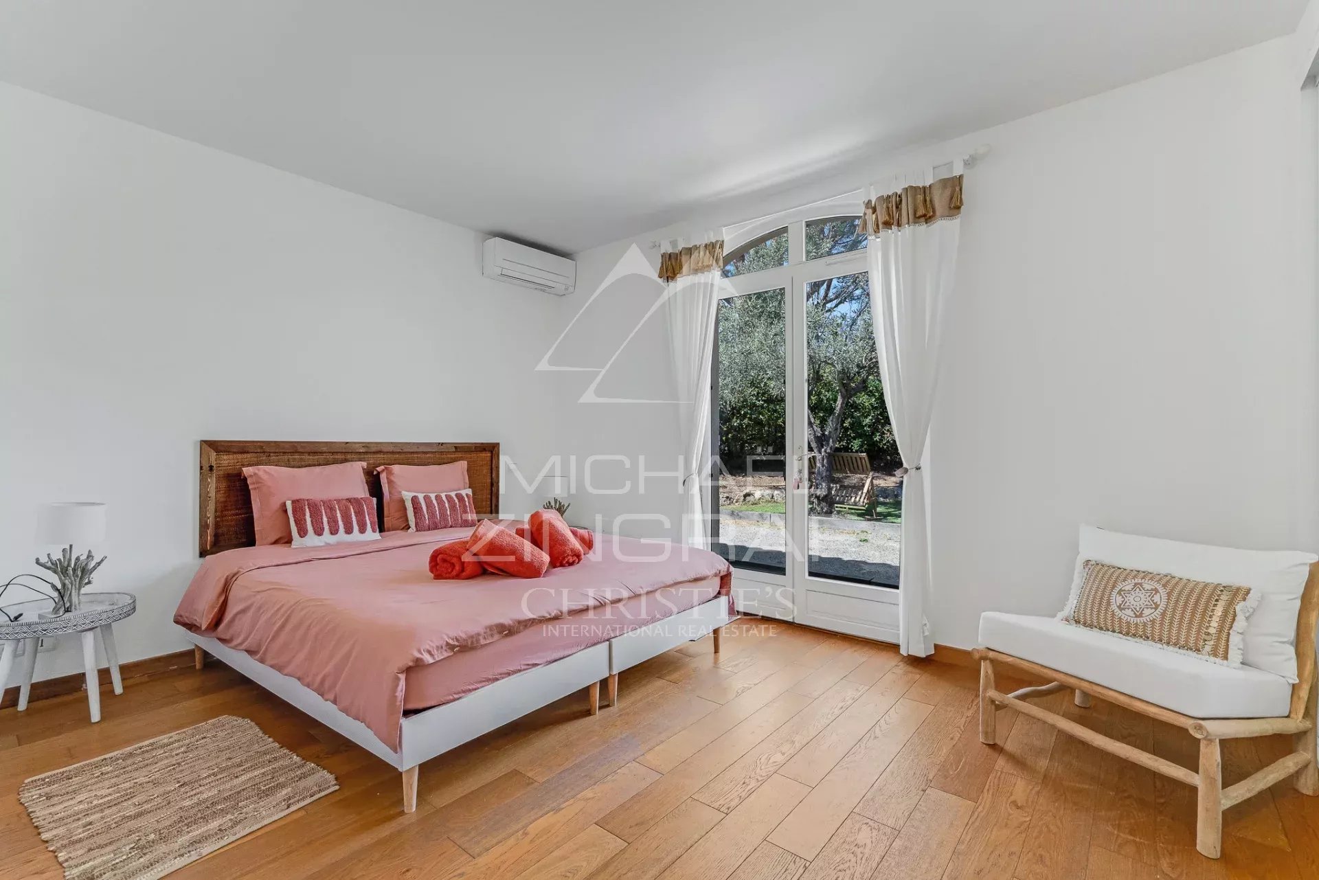 Bright bedroom with a pink-beddinged bed, wooden headboard, white walls, and glass doors opening to a garden outside.