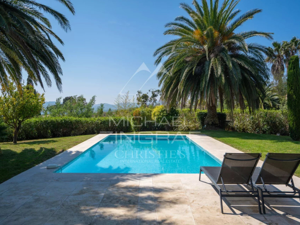 Rectangular swimming pool bordered by stone patio, with palm trees and green lawn in a sunny garden backdrop. Two lounge chairs sit at the edge.