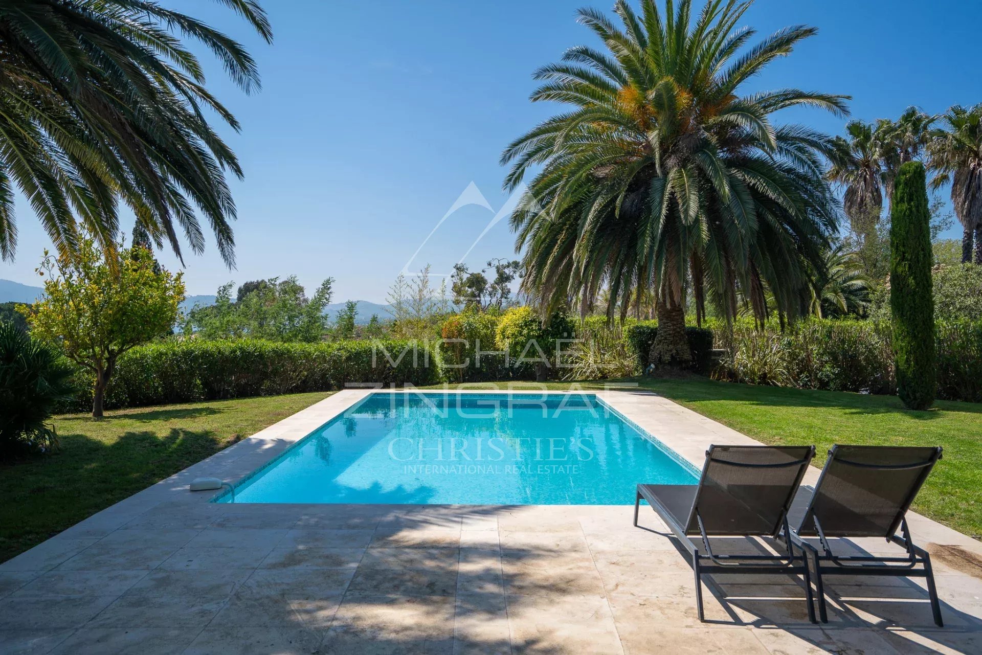 Rectangular swimming pool bordered by stone patio, with palm trees and green lawn in a sunny garden backdrop. Two lounge chairs sit at the edge.