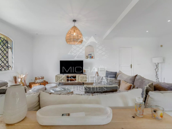 Bright, airy living room with white walls, a beige sectional sofa, and a low media console with a flat-screen TV. A woven pendant light hangs from the ceiling. Large window with a lattice outside adds natural light.