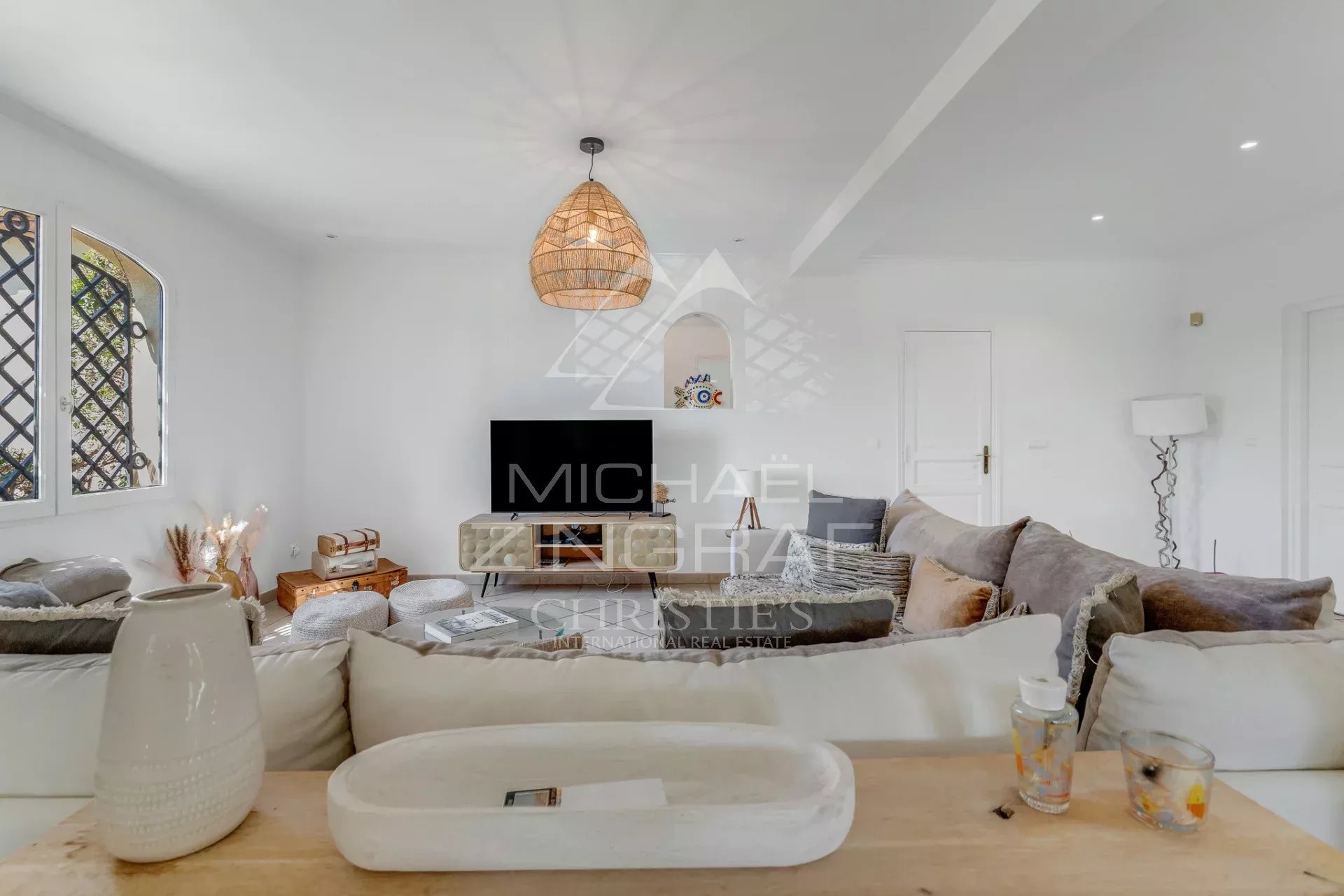 Bright, airy living room with white walls, a beige sectional sofa, and a low media console with a flat-screen TV. A woven pendant light hangs from the ceiling. Large window with a lattice outside adds natural light.