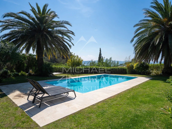 Rectangular pool with a stone deck, flanked by two palm trees and a green lawn, with the ocean visible in the distance.
