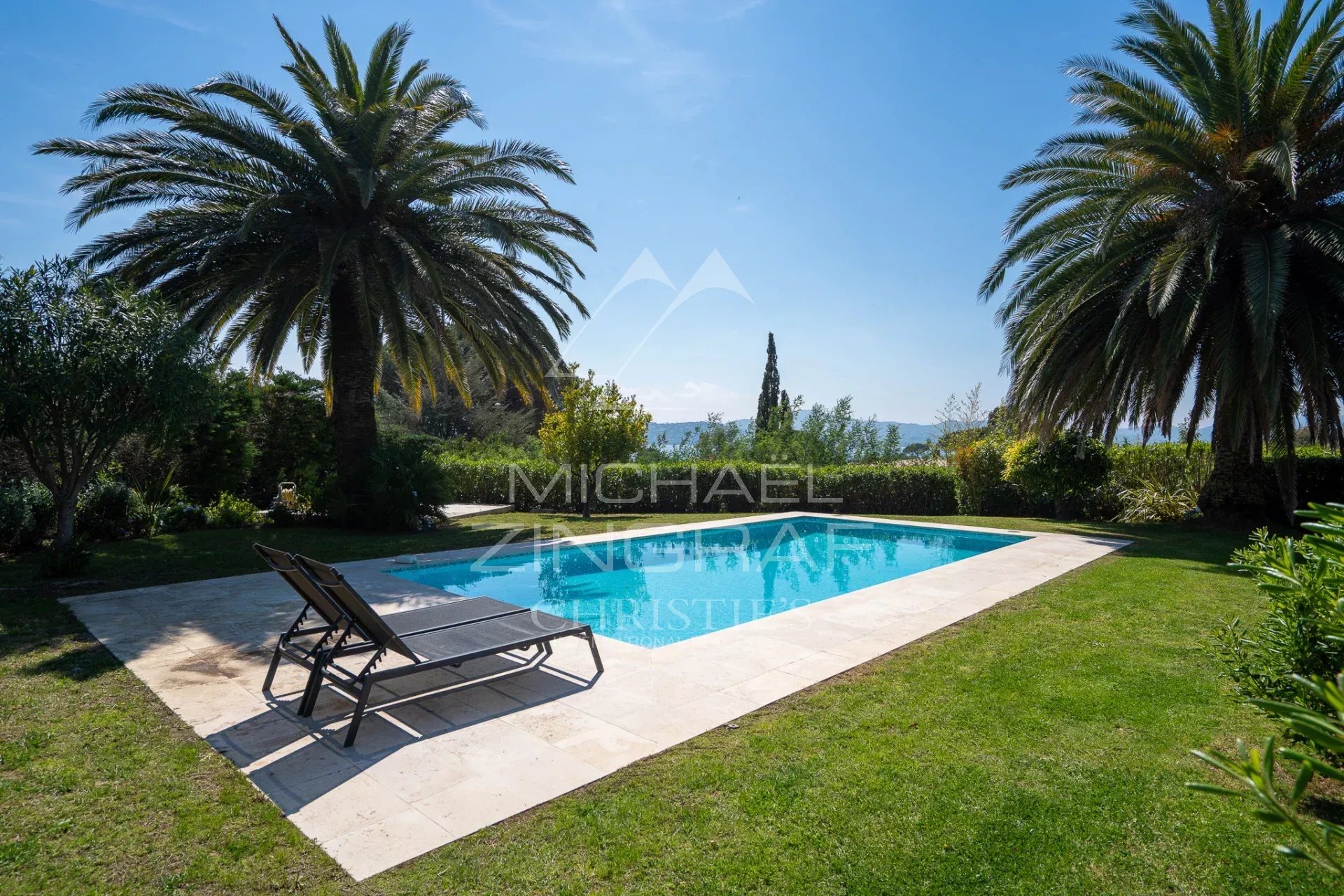 Rectangular pool with a stone deck, flanked by two palm trees and a green lawn, with the ocean visible in the distance.