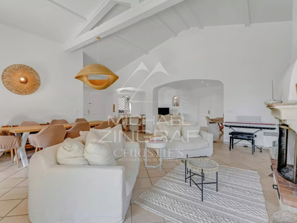 Bright open-plan living and dining room with white furniture, a wooden dining table, and a woven pendant light under a vaulted ceiling, near a fireplace on the right.