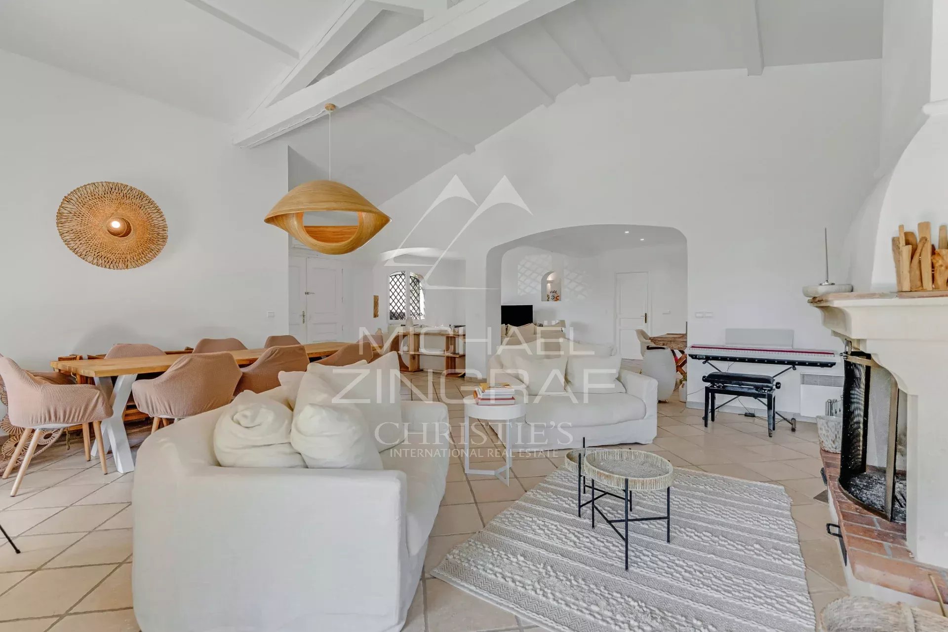 Bright open-plan living and dining room with white furniture, a wooden dining table, and a woven pendant light under a vaulted ceiling, near a fireplace on the right.