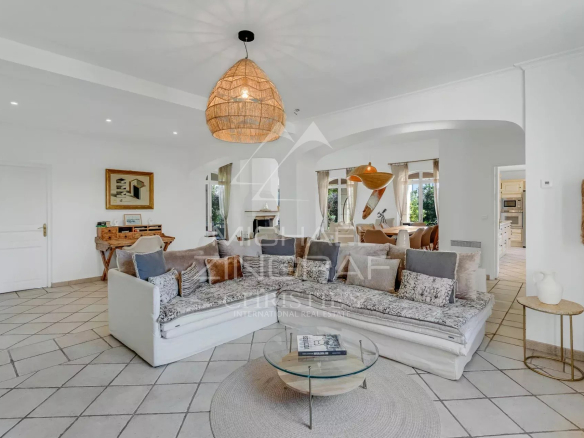 Bright open-plan living room with a white L-shaped sectional, patterned cushions, and a round glass coffee table under a woven pendant light.