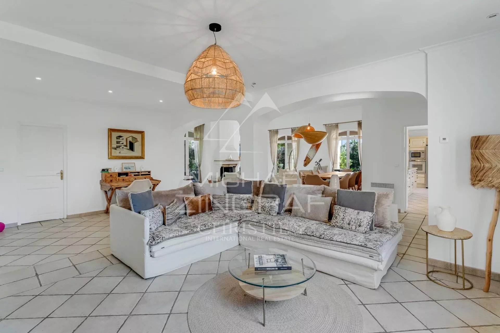 Bright open-plan living room with a white L-shaped sectional, patterned cushions, and a round glass coffee table under a woven pendant light.