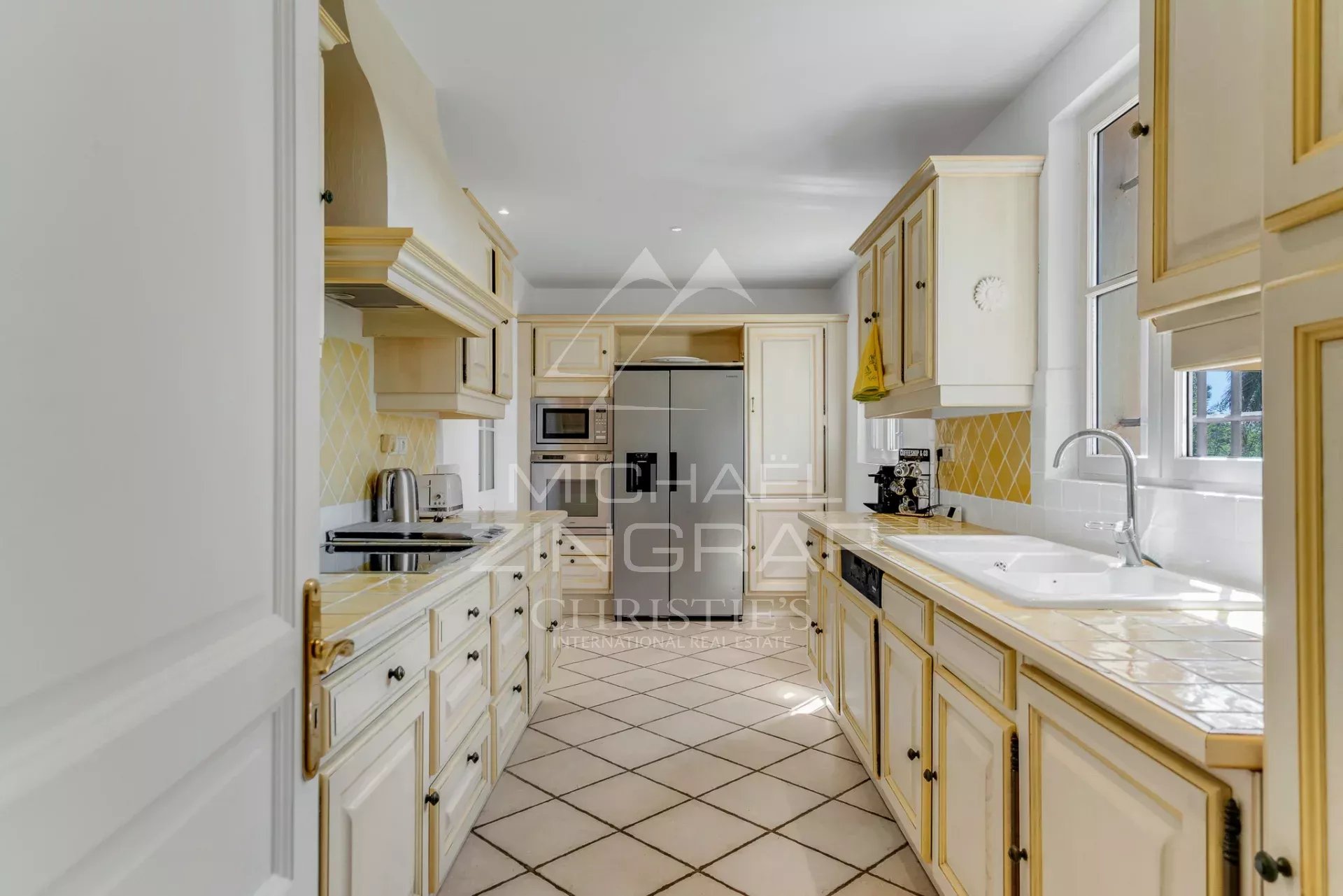 Bright cream kitchen with gold-trim cabinets, yellow tile backsplash, and stainless appliances along a narrow galley.