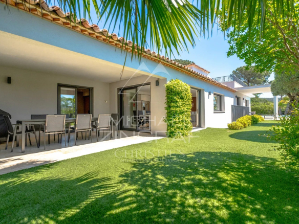 Outdoor patio of a white villa with a dining table, chairs, and a barbecue under a shaded veranda, plus a green lawn and palm fronds above.