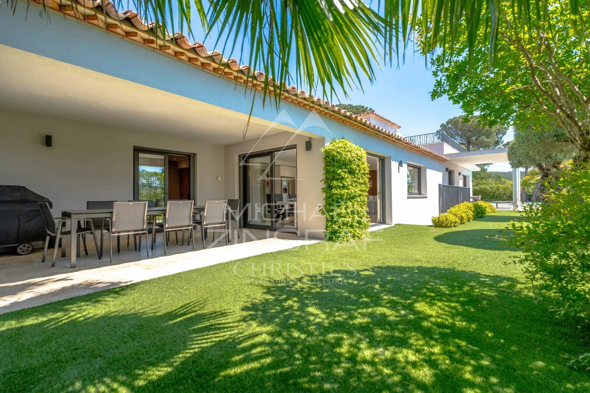 Outdoor patio of a white villa with a dining table, chairs, and a barbecue under a shaded veranda, plus a green lawn and palm fronds above.