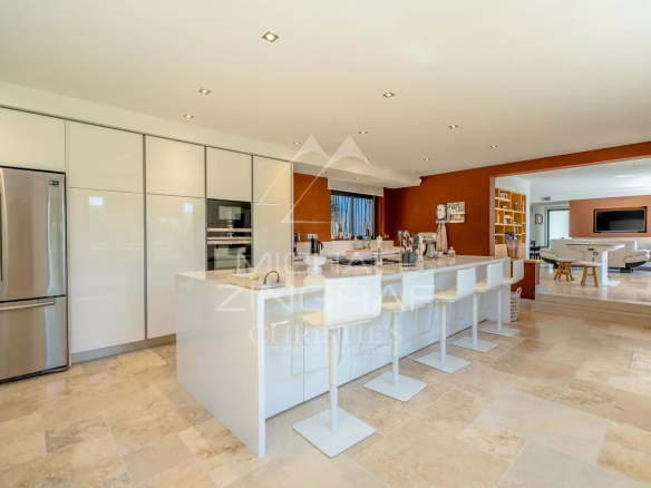 Open-plan kitchen with a white island, bar stools, and stainless steel fridge and ovens against glossy white cabinetry.