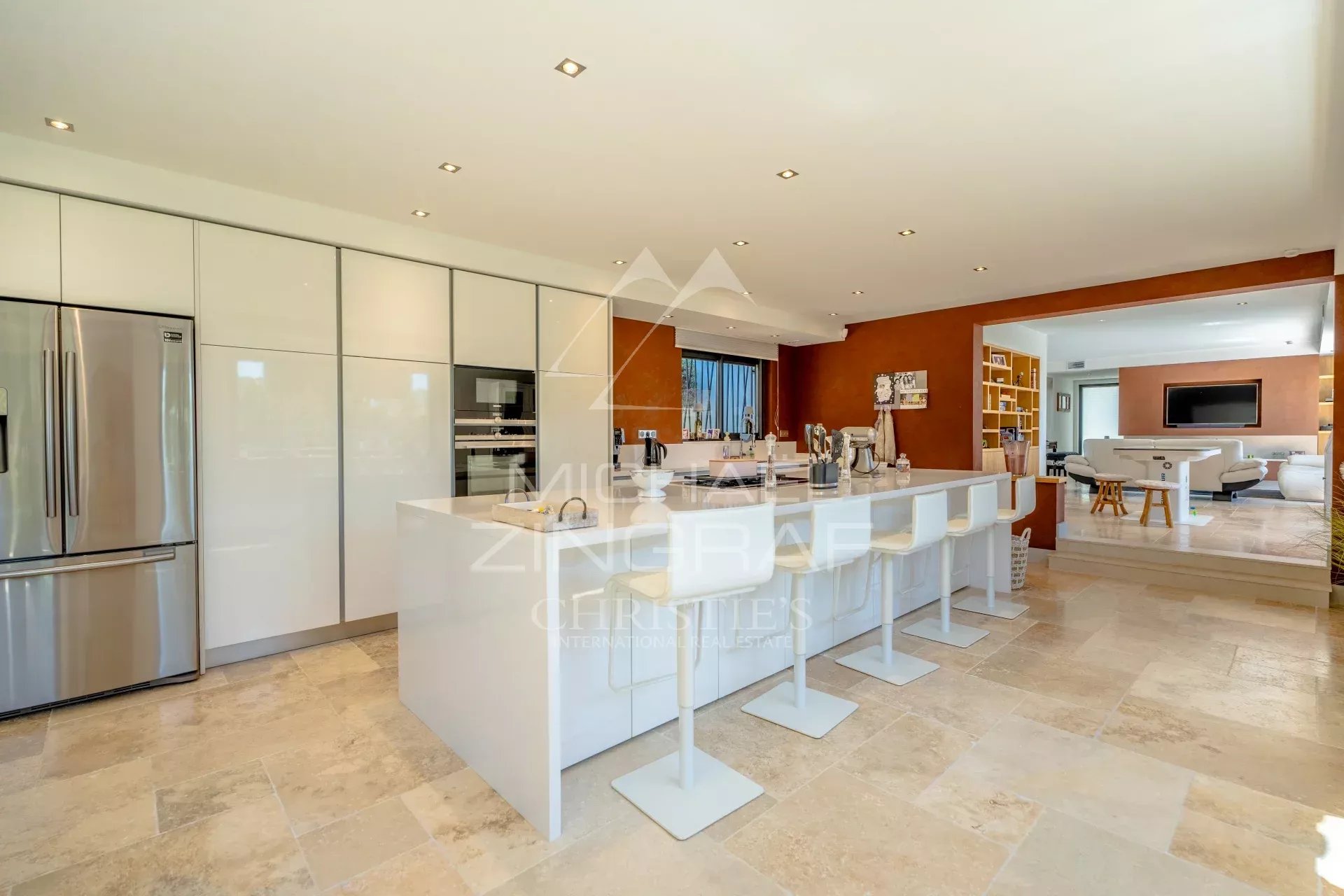 Open-plan kitchen with a white island, bar stools, and stainless steel fridge and ovens against glossy white cabinetry.