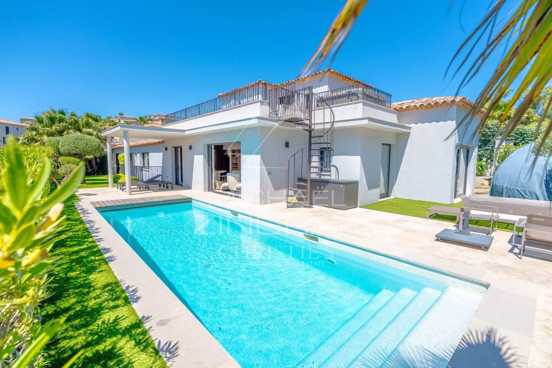 Modern white villa with a rectangular blue pool, spiral staircase, and outdoor lounge on a sunny day.