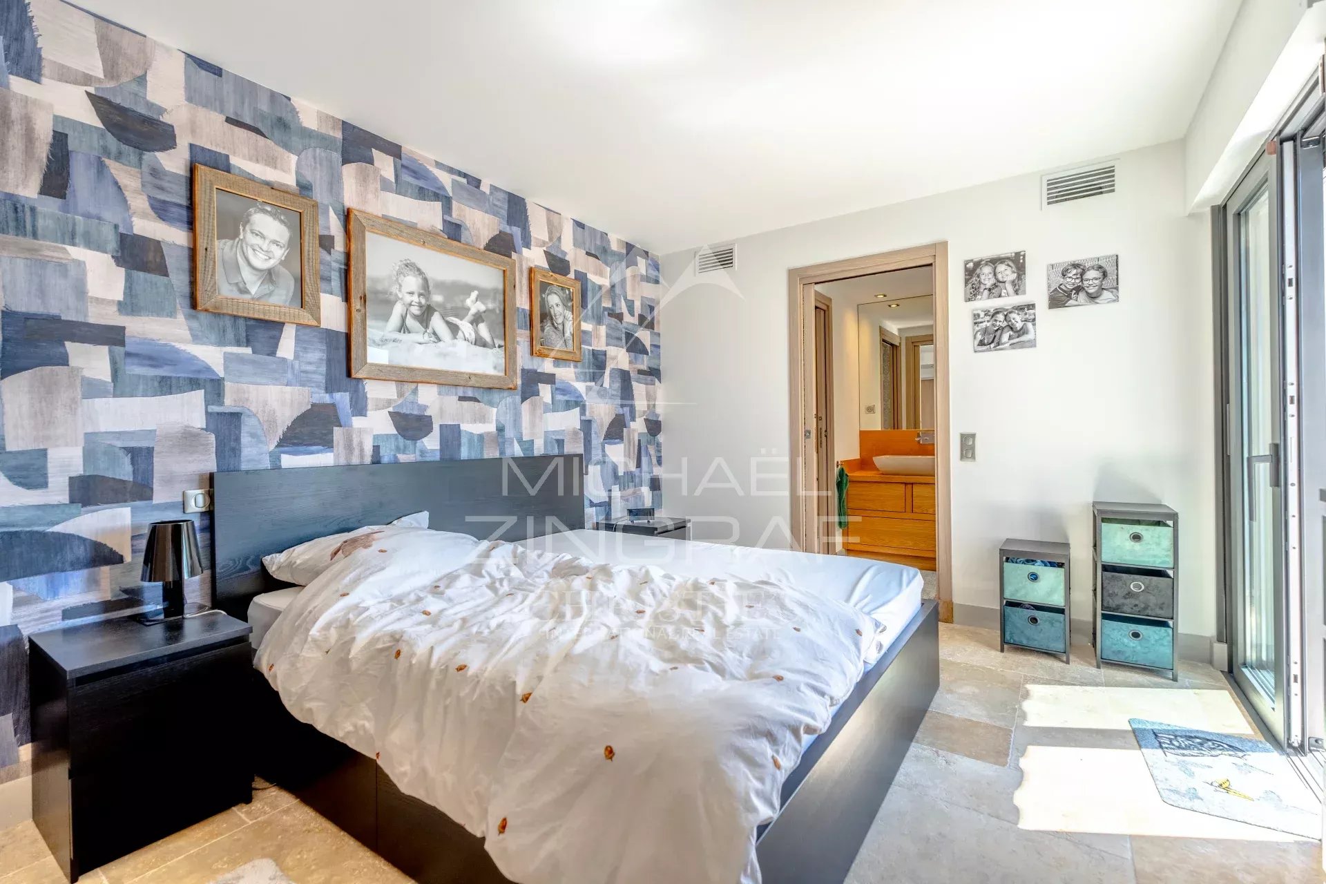 Bedroom with blue geometric wallpaper, a low dark bed, white duvet, and framed photos on the wall above the headboard.