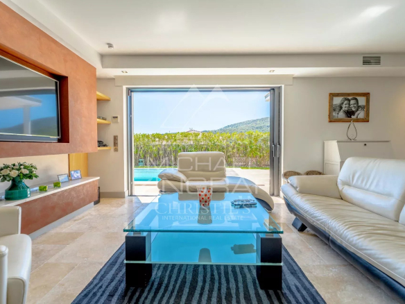Bright living room with white leather sofa, glass coffee table, and large sliding doors opening to a pool and green hedge beyond.
