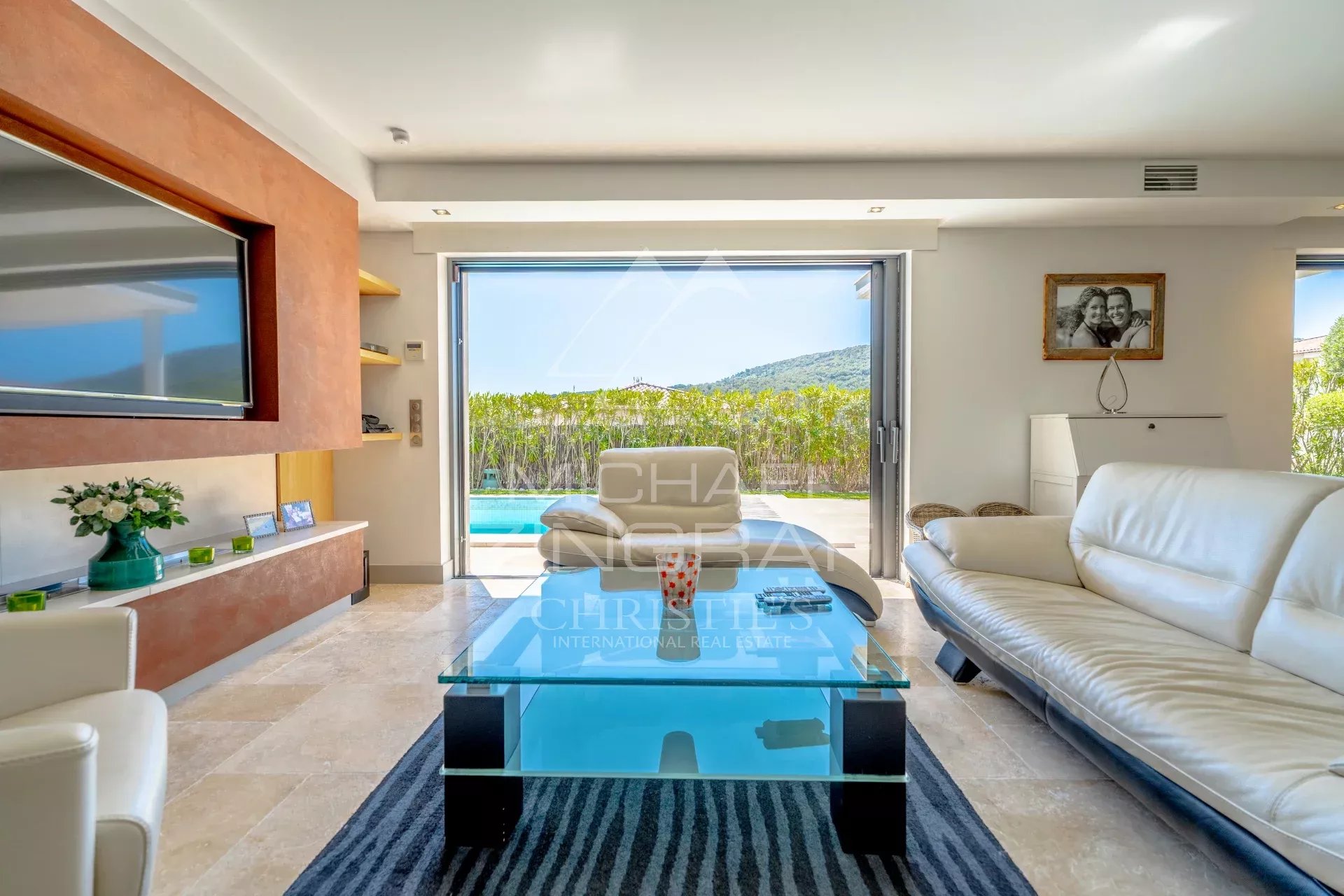 Bright living room with white leather sofa, glass coffee table, and large sliding doors opening to a pool and green hedge beyond.