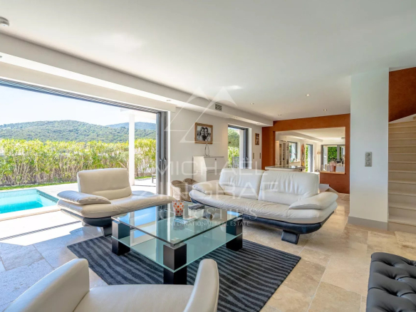 Open-plan living room with white leather sofas, glass coffee table, and a view to a pool and green hillside through large sliding doors.