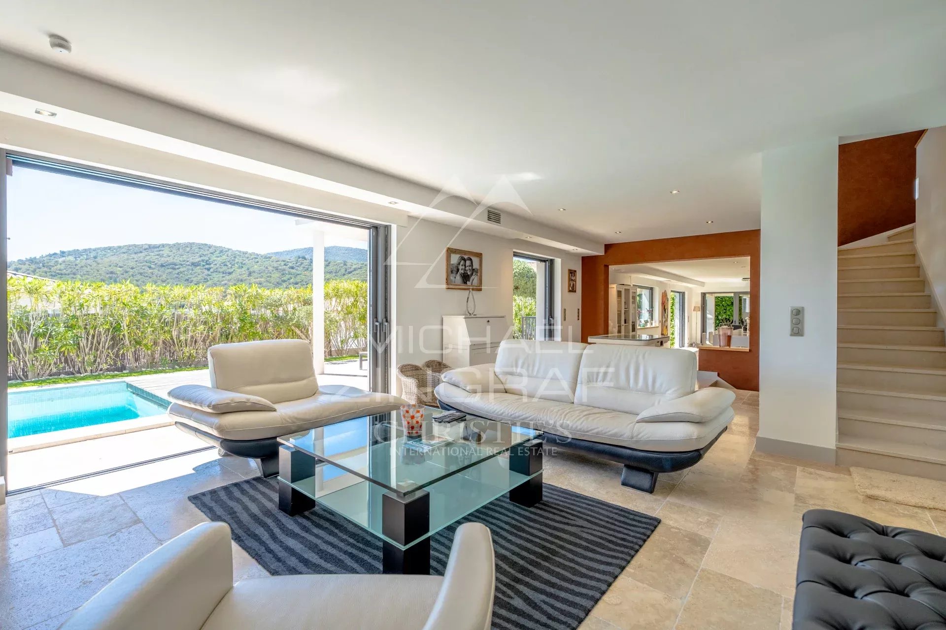 Open-plan living room with white leather sofas, glass coffee table, and a view to a pool and green hillside through large sliding doors.