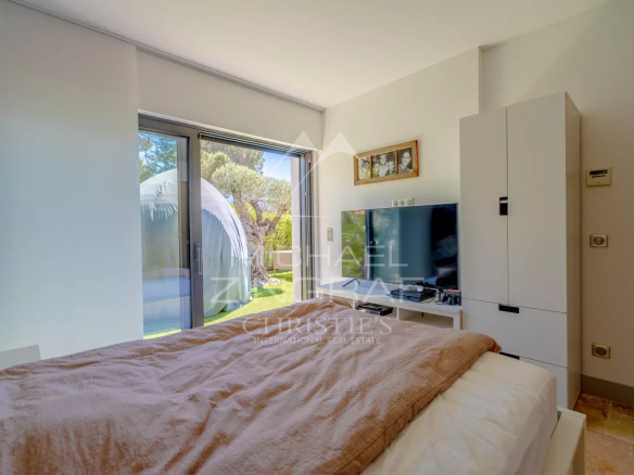 Bedroom with a brown blanket on the bed, white wardrobe, and a large glass door opening to a green garden outside.