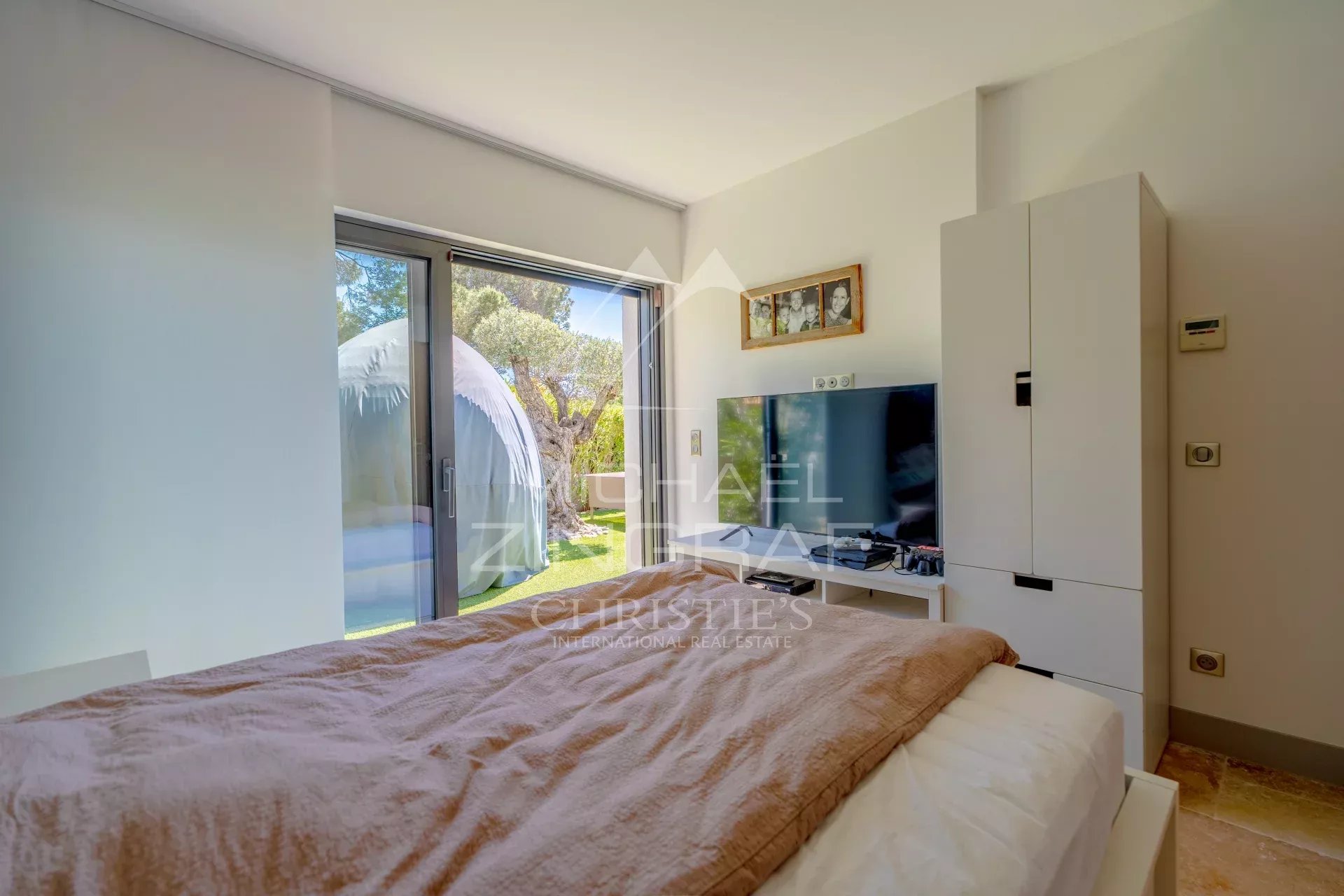Bedroom with a brown blanket on the bed, white wardrobe, and a large glass door opening to a green garden outside.