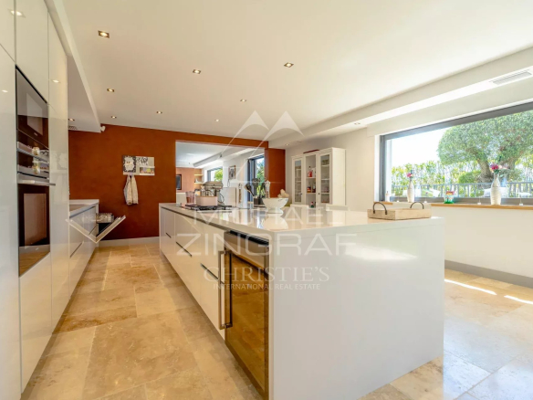 Bright open-plan kitchen with white glossy cabinets, a large central island, and a wide window overlooking greenery outside.