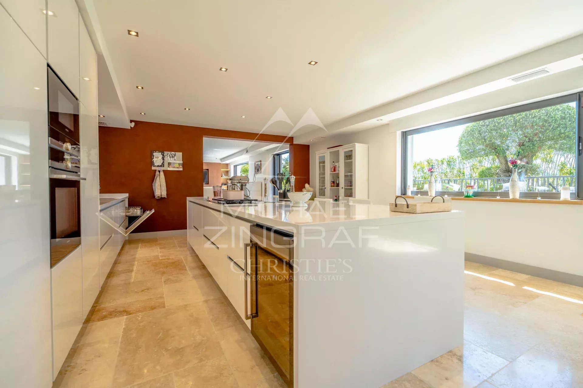 Bright open-plan kitchen with white glossy cabinets, a large central island, and a wide window overlooking greenery outside.