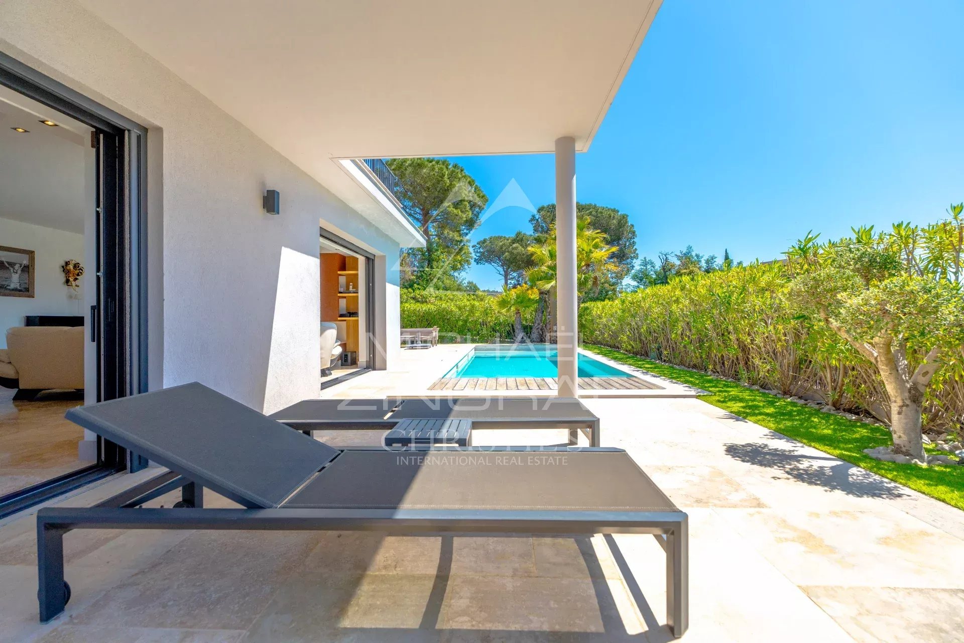 Outdoor patio with two black lounge chairs beside a pool, green hedges, and a bright blue sky beyond. Sliding glass door to a living room is open nearby.