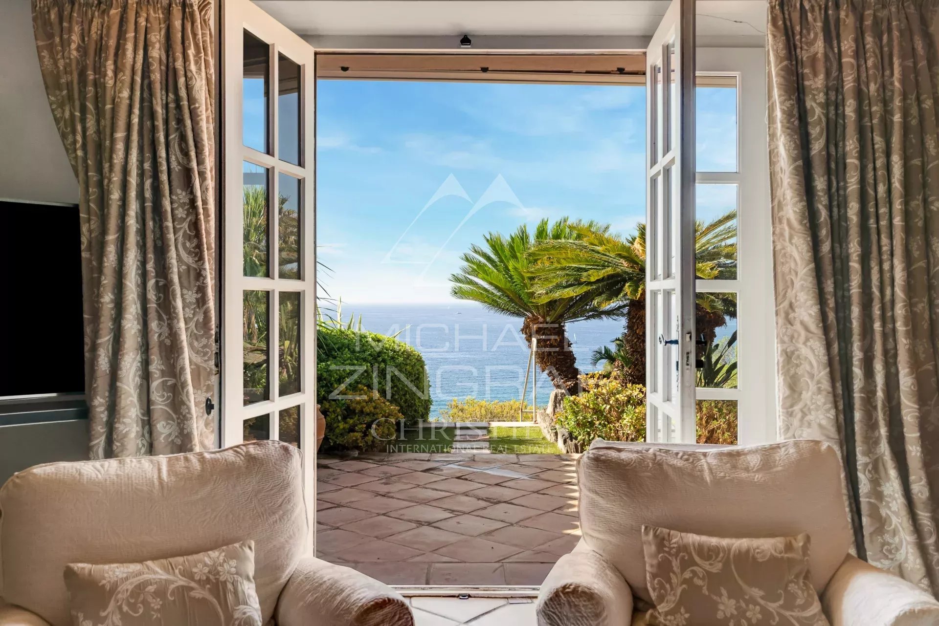 Vue depuis une villa avec des fauteuils, ouvrant sur un jardin et la mer.