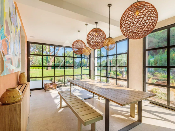 Salle à manger en véranda avec grandes fenêtres et mobilier en bois, vue sur le jardin.