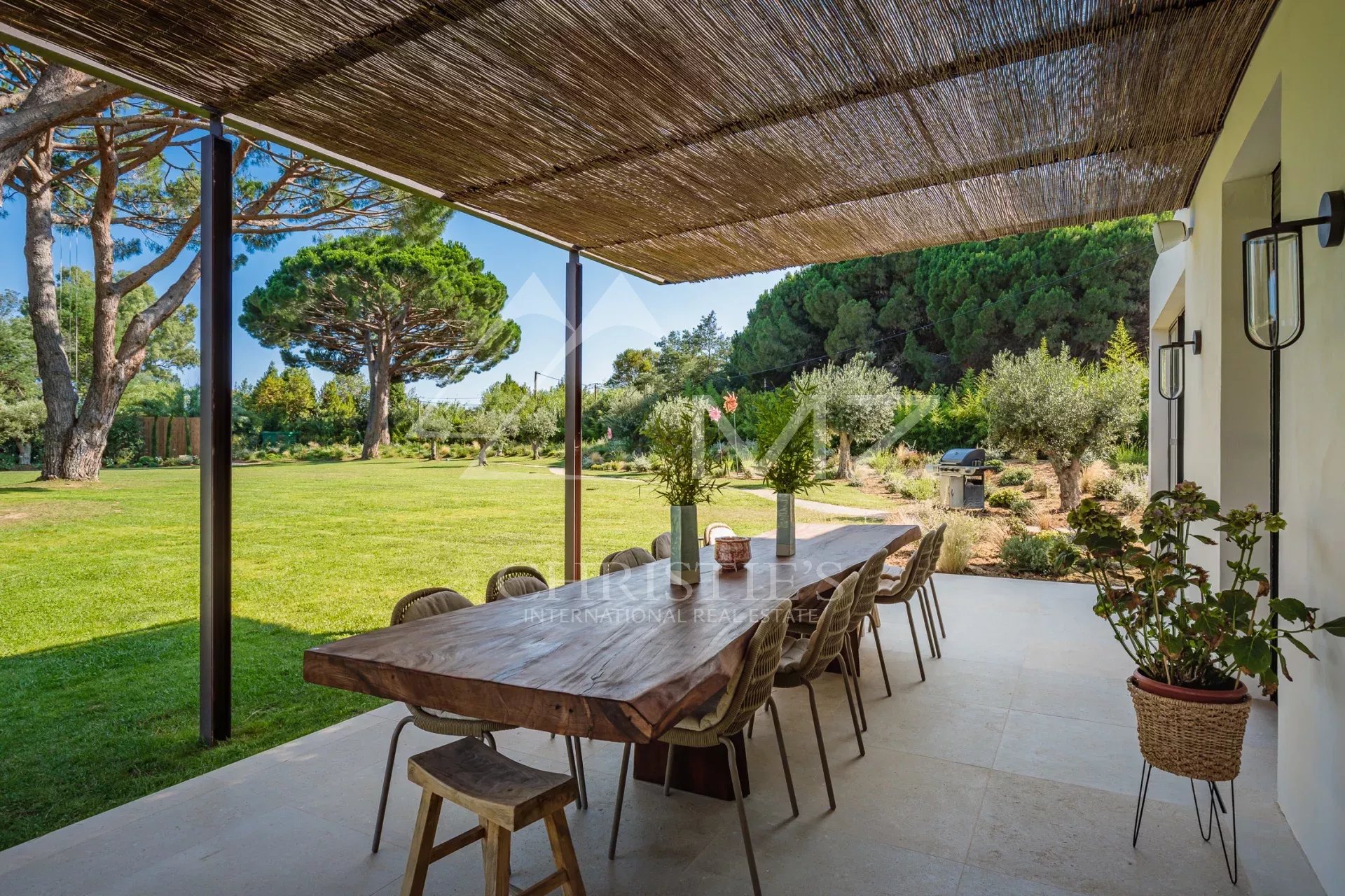 Terrasse de la Villa Shesh à Ramatuelle Terrasse d'une villa avec une grande table en bois, entourée d'un parc paysager.