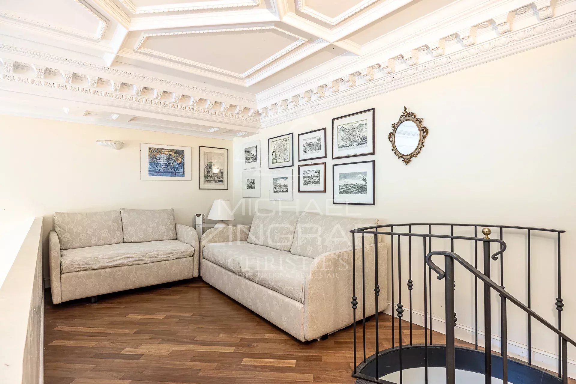 Cozy living corner with two beige patterned sofas, white ornate ceiling, and a wall of framed black-and-white art.