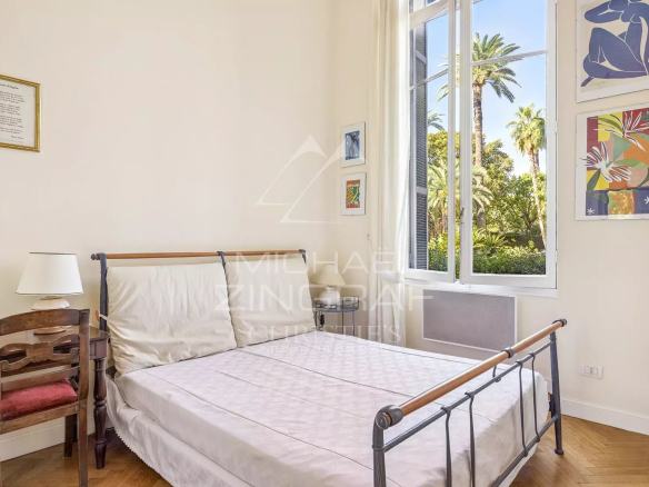 Bright bedroom with a metal-framed double bed, white bedding, and a large open window showing palm trees outside.