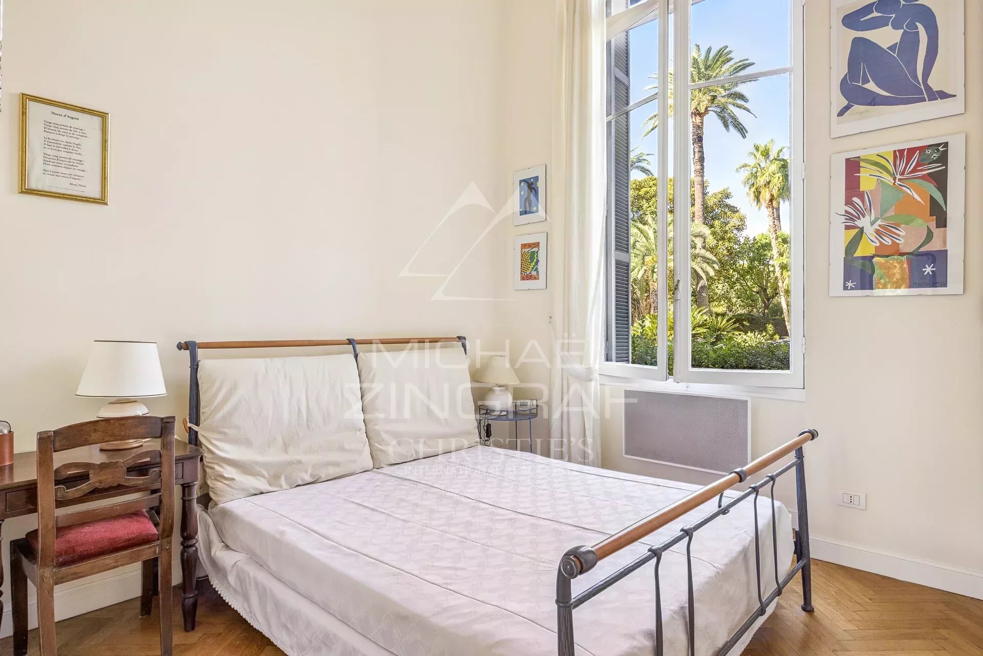 Bright bedroom with a metal-framed double bed, white bedding, and a large open window showing palm trees outside.
