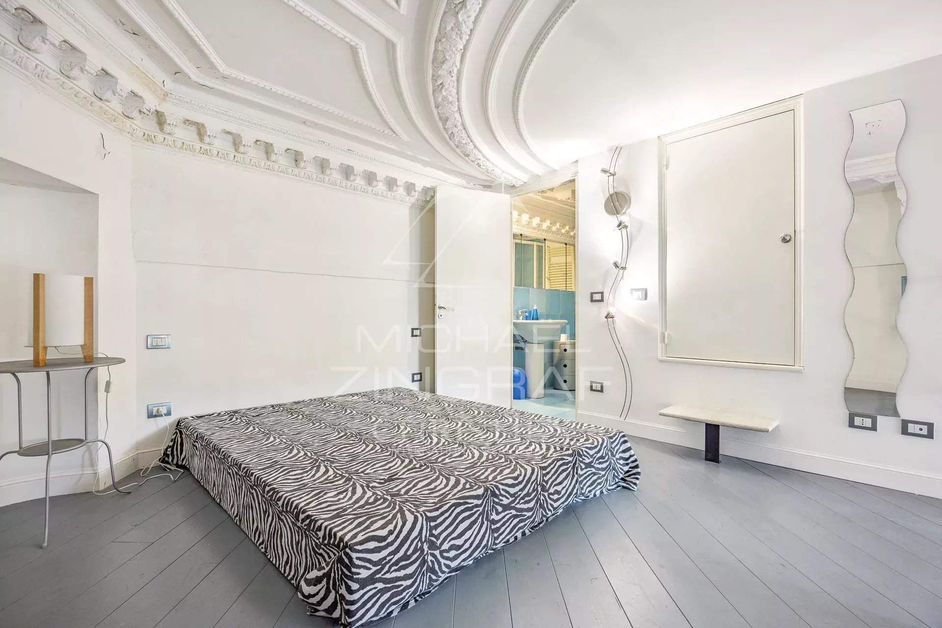Bedroom with a zebra-patterned bedspread, ornate white ceiling molding, and an open door to a blue-tiled bathroom at the side.